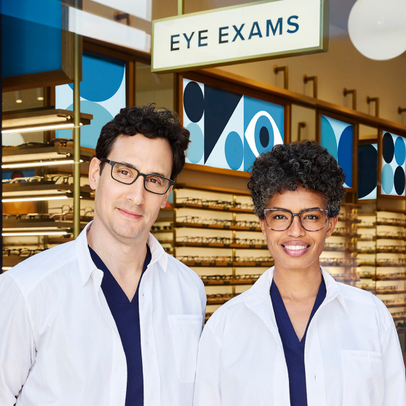 Two optometrists smiling in front of an eye exam sign
